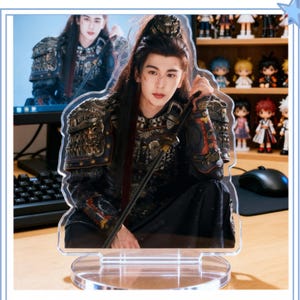 May include: A clear acrylic standee featuring a printed image of a person with long dark hair dressed in ornate dark armor and holding a sword. The figure is depicted in a kneeling pose, mounted on a transparent circular base for display purposes.