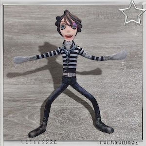 May include: A fabric doll of a character with brown hair, a striped shirt, and dark blue pants. The doll has outstretched arms and is standing on a gray surface. The doll has a detailed face with a smiling expression.