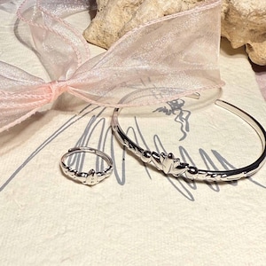 Könnte beinhalten: Ein silbernes Ring- und Armband-Set mit floralem Design. Das Armband ist im Manschettenstil. Ein rosa Band befindet sich im Hintergrund. Der Schmuck liegt auf einer cremefarbenen Oberfläche.