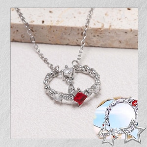 Puede incluir: Collar plateado con un colgante circular con piedras preciosas claras y rojas. El colgante tiene un diseño en forma de estrella con una gema roja cuadrada en el centro. El collar está sobre una superficie blanca.