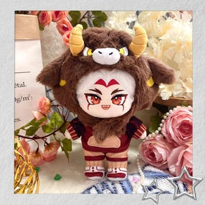 May include: A plush doll dressed in a brown bull-themed hood and outfit. The doll features a white face with red details, large red eyes, and a small smile. The outfit includes a maroon top and tan trousers with red and white striped accents.