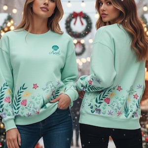 May include: Two mint green sweatshirts with floral and seashell designs. The sweatshirts have the word "Ariel" written on the front. The sleeves and lower portion of the sweatshirts are decorated with colorful flowers and sea elements. The models are wearing blue jeans and black pants.