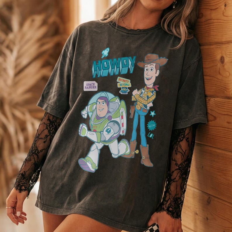 Peut inclure: T-shirt gris fonc&eacute; avec Buzz l'&Eacute;clair et Woody de Toy Story. Le t-shirt porte le mot "HOWDY" en bleu et les phrases "SPACE RANGER" et "INFINITY BEYOND". Le t-shirt a des manches courtes et est port&eacute; avec une manche en dentelle noire.