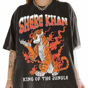 Puede incluir: Camiseta negra con un diseño de Shere Khan, un tigre rodeado de llamas. El texto "SHERE KHAN" está encima del tigre y "KING OF THE JUNGLE" debajo. El diseño es naranja y blanco.
