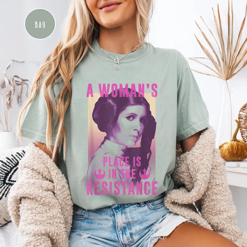 May include: A light blue t-shirt featuring a pink graphic of Princess Leia from Star Wars. The graphic includes the text "A Woman's Place is in the Resistance".