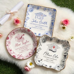 May include: Three decorative ceramic plates with floral and graduation cap designs. Each plate features a name, university, and graduation year. The plates are white with gold rims and are surrounded by flowers and a rolled-up diploma.