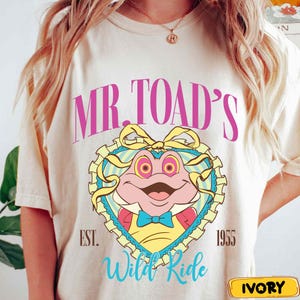 May include: Ivory-colored t-shirt featuring a cartoon frog design. The design includes the text "MR. TOAD'S" in pink, a heart-shaped frame, and the words "Wild Ride" in blue. The shirt also has the text "EST. 1955".