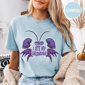 Puede incluir: Una camiseta azul claro con una ilustraci&oacute;n de cangrejo de dibujos animados morado y el texto "Me com&iacute; a mi abuela".