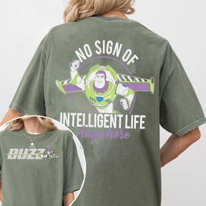 May include: Olive green t-shirt featuring a graphic of Buzz Lightyear with the text "NO SIGN OF INTELLIGENT LIFE Anywhere". The front of the shirt has the word "BUZZ" in silver block letters and a small graphic of Buzz Lightyear.