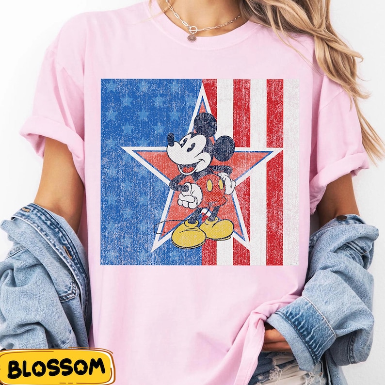 Pu&ograve; includere: T-shirt rosa chiaro con una grafica invecchiata di Mickey Mouse all'interno di una stella rossa, su un campo blu di stelle e strisce rosse e bianche. Il design evoca il tema della bandiera americana vintage.