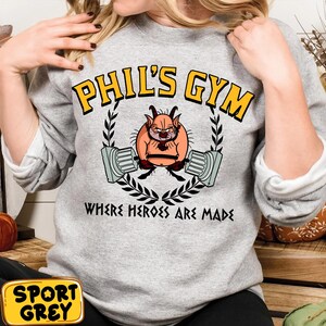 May include: A heather grey sweatshirt with the text "PHIL'S GYM" in yellow block letters. The design features a cartoon character lifting weights, surrounded by a laurel wreath, with the words "WHERE HEROES ARE MADE" below.