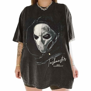 Könnte beinhalten: Dunkelgraues T-Shirt mit einer Grafik von Taskmasters Maske und Kapuze. Das Design ist in Weiß und Grau gehalten, mit dem Text "Taskmaster" und "Thunderbolts" in einer Schriftart. Das Shirt hat kurze Ärmel und eine lockere Passform.