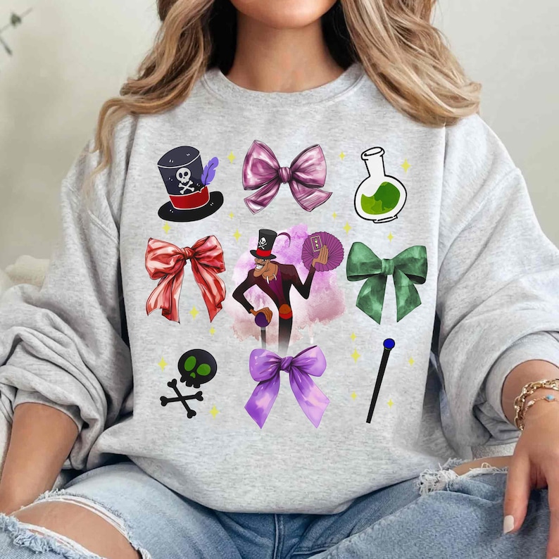 May include: A light gray sweatshirt featuring various cartoon graphics. The graphics include a top hat with a skull, bows in pink, red, and green, a potion bottle, a skull and crossbones, a magician character, and a magic wand.