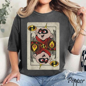 Può includere: Una maglietta nera con un'illustrazione in stile vintage di un personaggio dei cartoni animati con un abito rosso e nero che tiene una carta da gioco. La carta ha un cuore rosso e il testo "Jack" su di essa. Il personaggio indossa una maschera rossa e un mantello rosso.