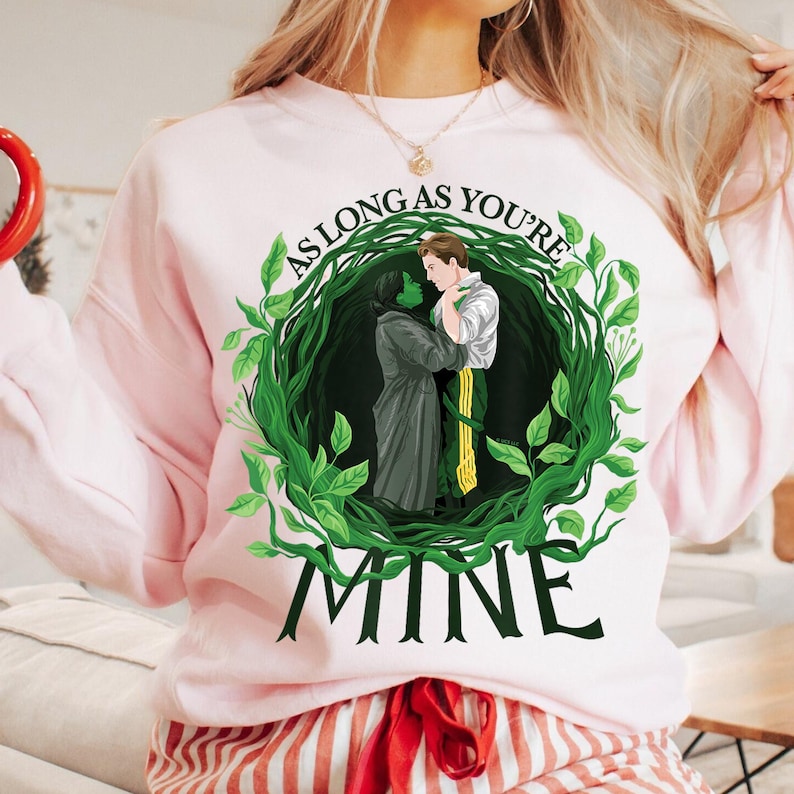 May include: A pale pink sweatshirt with a green and white illustration of two figures embracing within a leafy wreath. The text "AS LONG AS YOU'RE" arches above the illustration, with "MINE" below. The sweatshirt has long sleeves.