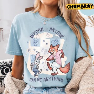 Pode incluir: Camiseta azul claro com uma ilustra&ccedil;&atilde;o de desenho animado de uma raposa e um coelho, com o texto "WHERE ANYONE CAN BE ANYTHING". O design inclui pegadas e uma palmeira. A camiseta &eacute; de gola redonda.