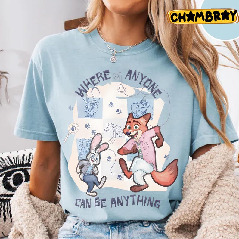 Pode incluir: Camiseta azul claro com uma ilustra&ccedil;&atilde;o de desenho animado de uma raposa e um coelho, com o texto "WHERE ANYONE CAN BE ANYTHING". O design inclui pegadas e uma palmeira. A camiseta &eacute; de gola redonda.
