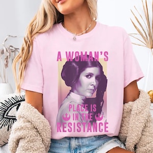 May include: Pink t-shirt featuring a graphic of Princess Leia from Star Wars. The graphic reads "A Woman's Place Is In The Resistance".