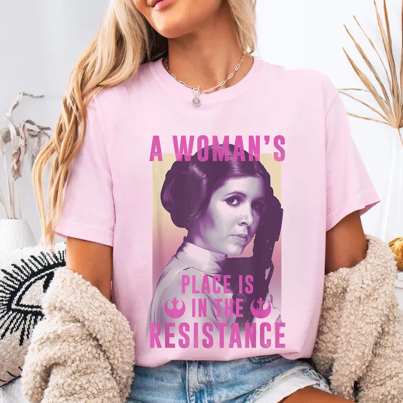 May include: Pink t-shirt featuring a graphic of Princess Leia from Star Wars. The graphic reads "A Woman's Place Is In The Resistance".