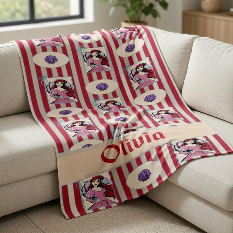 Op de afbeelding: Een rood-wit gestreepte deken met een cartoon prinsessenontwerp. De deken heeft een cr&egrave;mekleurige rand met de naam "Olivia" in het rood gedrukt. Het ontwerp bevat afbeeldingen van een prinses en zeeschelpen.