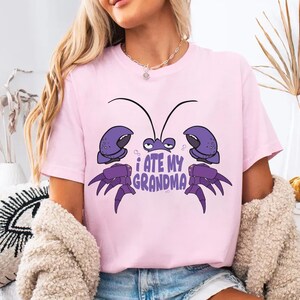 Puede incluir: Camiseta rosa con un cangrejo de dibujos animados morado y el texto "Me com&iacute; a mi abuela".