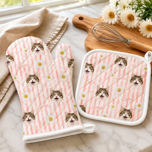 May include: A quilted oven mitt and square pot holder set featuring a repeating pattern of white daisies, small hearts, and the face of a tabby cat with the name Luna written in cursive script against a background of vertical light pink stripes.