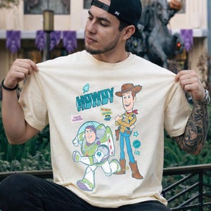 Peut inclure: T-shirt cr&egrave;me avec les personnages de dessins anim&eacute;s Buzz l'&Eacute;clair et Woody de Toy Story. Le t-shirt porte le mot "HOWDY" en turquoise, ainsi que d'autres graphiques et du texte. Le t-shirt est tenu par une personne.
