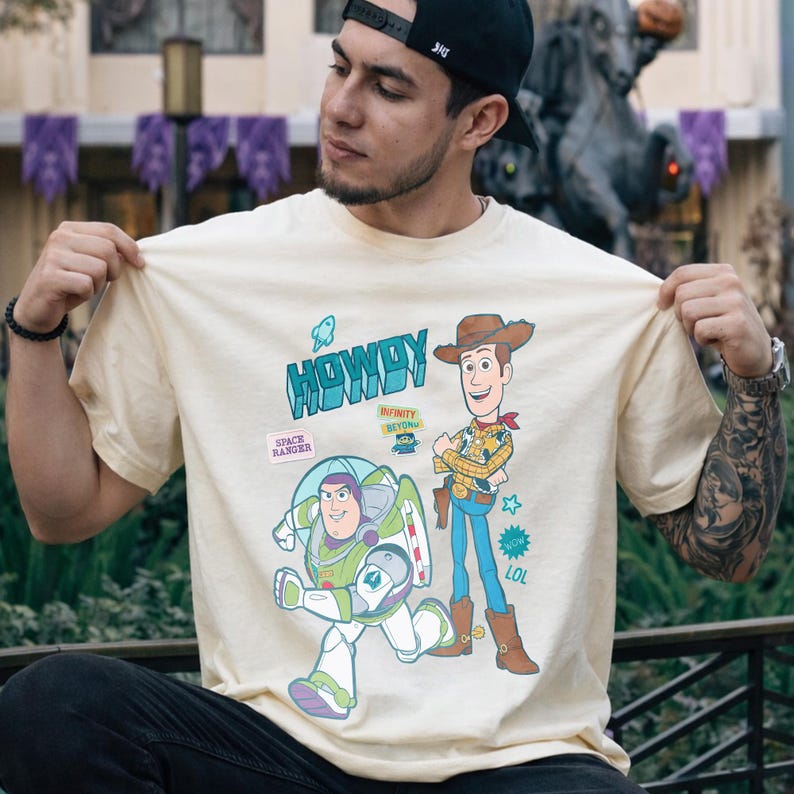 Peut inclure: T-shirt cr&egrave;me avec les personnages de dessins anim&eacute;s Buzz l'&Eacute;clair et Woody de Toy Story. Le t-shirt porte le mot "HOWDY" en turquoise, ainsi que d'autres graphiques et du texte. Le t-shirt est tenu par une personne.