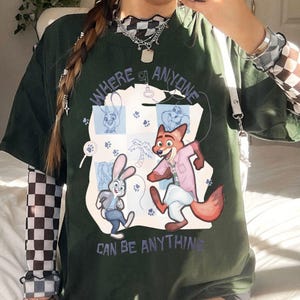 Pode incluir: T-shirt verde escuro com os personagens de desenho animado Judy Hopps e Nick Wilde de Zootopia. O texto "WHERE ANYONE CAN BE ANYTHING" &eacute; roxo claro. O design inclui pegadas e uma palmeira.