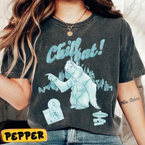 Puede incluir: Camiseta gris oscuro con un diseño azul claro de un perezoso con traje y el texto "Chillout!". El diseño incluye un paisaje urbano y la palabra "Zootropolis". La camiseta tiene un aspecto vintage y desgastado.
