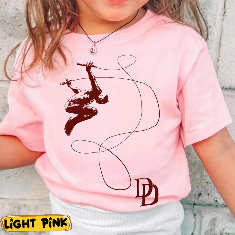 May include: Light pink t-shirt featuring a brown graphic of a figure swinging on a rope, with the initials "DD" below. The shirt has short sleeves and a crew neck. The words "LIGHT PINK" are in a yellow box at the bottom.