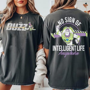 May include: Dark gray t-shirts featuring Buzz Lightyear from Pixar's Toy Story. One shirt has "Buzz" in white and yellow, with Buzz Lightyear. The other shirt has Buzz Lightyear with the text "NO SIGN OF INTELLIGENT LIFE Anywhere".