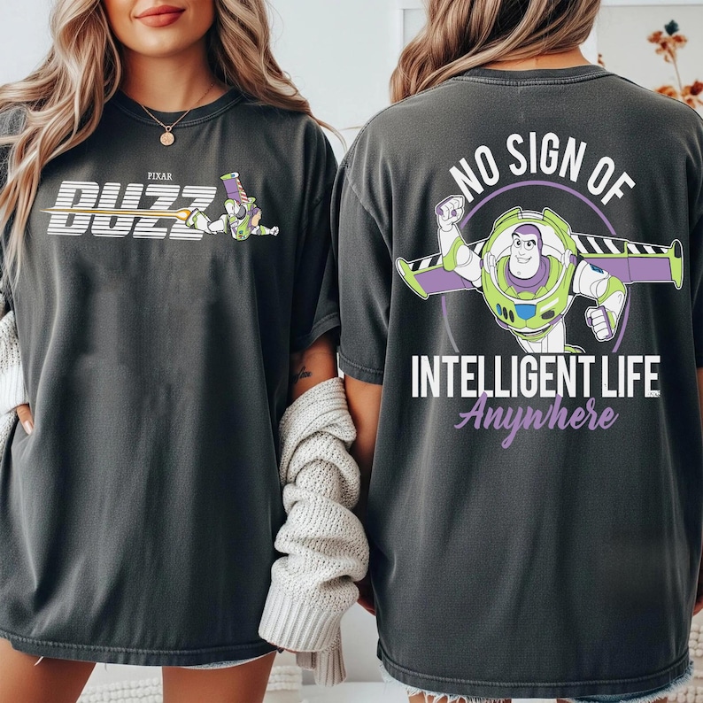 May include: Dark gray t-shirts featuring Buzz Lightyear from Pixar's Toy Story. One shirt has "Buzz" in white and yellow, with Buzz Lightyear. The other shirt has Buzz Lightyear with the text "NO SIGN OF INTELLIGENT LIFE Anywhere".