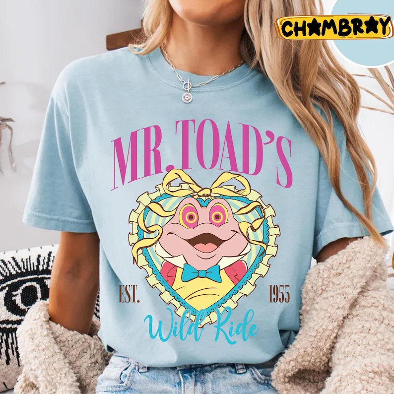 May include: Light blue t-shirt featuring a cartoon image of Mr. Toad inside a heart-shaped frame. The design includes the text "MR. TOAD'S Wild Ride" and the year "1955". The shirt has a crew neck and short sleeves.