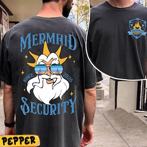 May include: Dark gray t-shirt with a graphic of a cartoon character wearing sunglasses, with the text "MERMAID SECURITY SINCE 2025". The shirt also features a smaller graphic on the upper right chest with the words "MERMAID SECURITY".