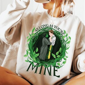 May include: A cream-coloured sweatshirt with a green and black illustration of two figures embracing within a leafy wreath. The text "AS LONG AS YOU'RE" arches above the image, with "MINE" below.