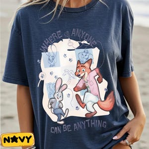 Pode incluir: Camiseta azul marinho com uma ilustra&ccedil;&atilde;o de desenho animado de uma raposa e um coelho, com o texto "WHERE ANYONE CAN BE ANYTHING". O design inclui pegadas e outros elementos de desenho animado. Uma etiqueta amarela com a palavra "NAVY" est&aacute; no canto inferior esquerdo.