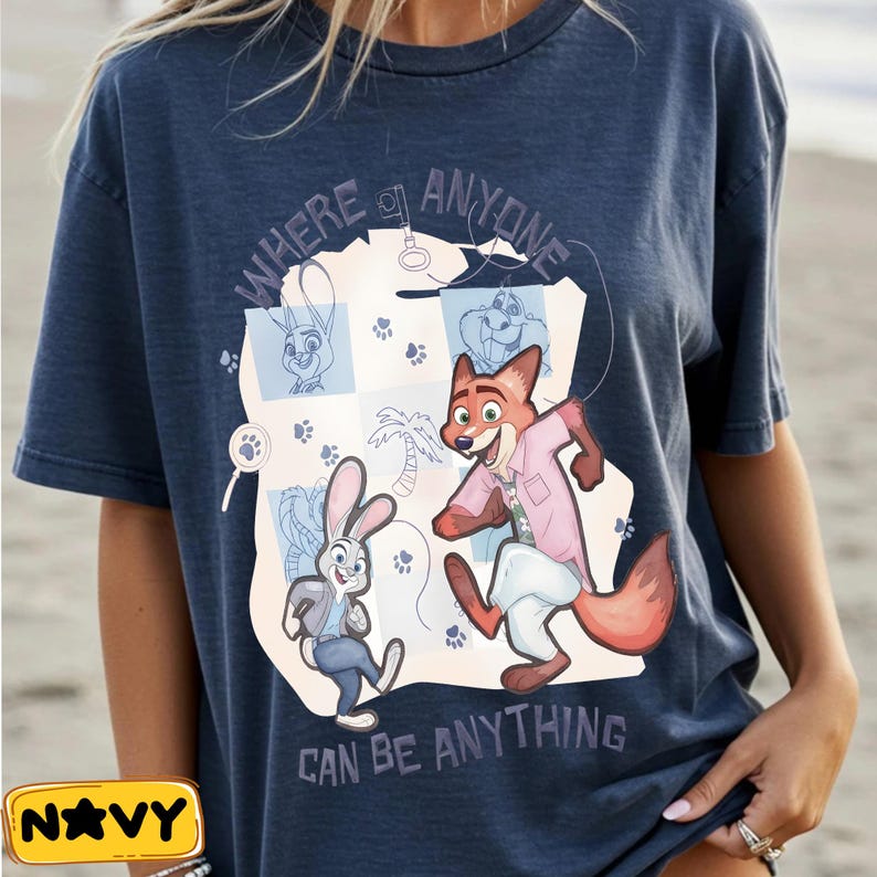 Pode incluir: Camiseta azul marinho com uma ilustra&ccedil;&atilde;o de desenho animado de uma raposa e um coelho, com o texto "WHERE ANYONE CAN BE ANYTHING". O design inclui pegadas e outros elementos de desenho animado. Uma etiqueta amarela com a palavra "NAVY" est&aacute; no canto inferior esquerdo.