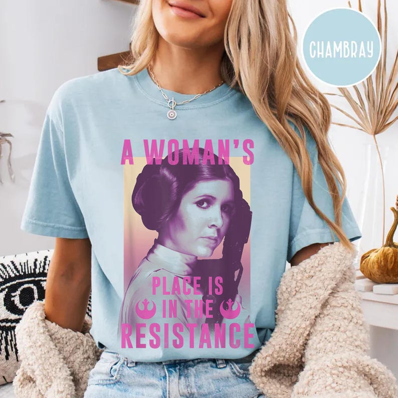 May include: A light blue t-shirt featuring a pink graphic of Princess Leia from Star Wars. The graphic includes the text "A Woman's Place is in the Resistance".