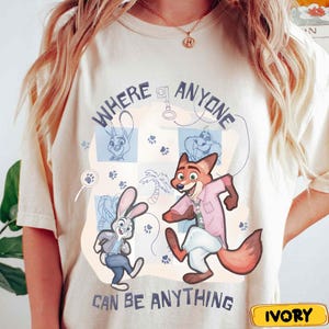 Pode incluir: Camiseta cor marfim com personagens de desenhos animados e o texto "WHERE ANYONE CAN BE ANYTHING". O design inclui ilustra&ccedil;&otilde;es de um coelho e uma raposa, juntamente com pegadas e outros gr&aacute;ficos. A camisa tem gola redonda e mangas curtas.