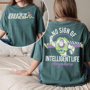 May include: Teal green t-shirt featuring Buzz Lightyear from Pixar's Toy Story. The front of the shirt displays "Buzz" with an image of Buzz Lightyear. The back of the shirt has the text "NO SIGN OF INTELLIGENT LIFE Anywhere" with a graphic of Buzz Lightyear.