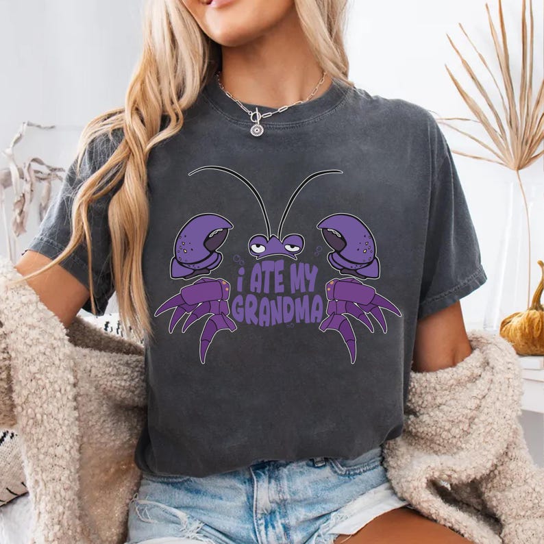 Puede incluir: Una camiseta gris oscuro con un cangrejo de dibujos animados morado y el texto "Me com&iacute; a mi abuela".
