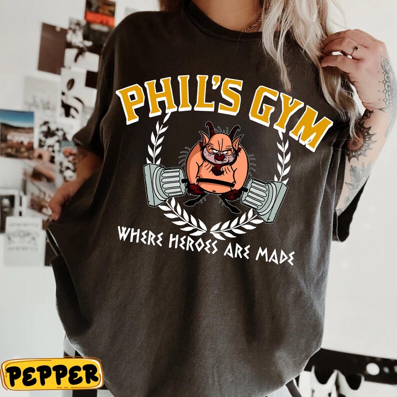 May include: Dark gray t-shirt with the text "PHIL'S GYM" in yellow, above a cartoon character lifting weights. The phrase "WHERE HEROES ARE MADE" is below the graphic. The design is framed by a white laurel wreath.