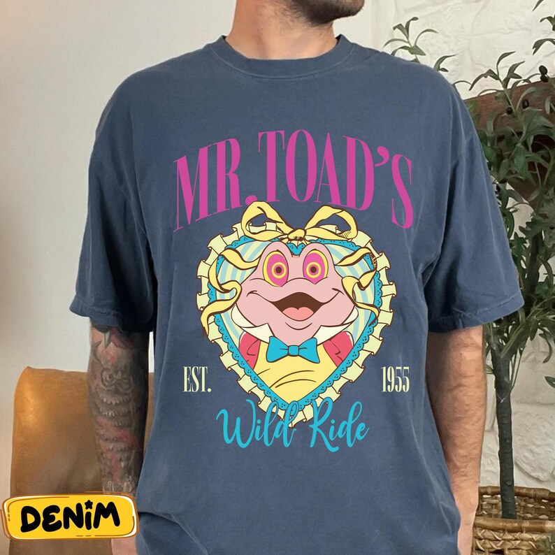 May include: A navy blue t-shirt featuring a cartoon image of Mr. Toad inside a heart-shaped frame. The text "MR. TOAD'S" is arched above the image in pink, with "EST. 1955" and "Wild Ride" below in teal.