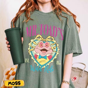 May include: Olive green t-shirt featuring a cartoon frog design with the text "Mr. Toad's Wild Ride" and the year "1955". A dark green studded tumbler is held in the left hand. The word "MOSS" is on a yellow label.