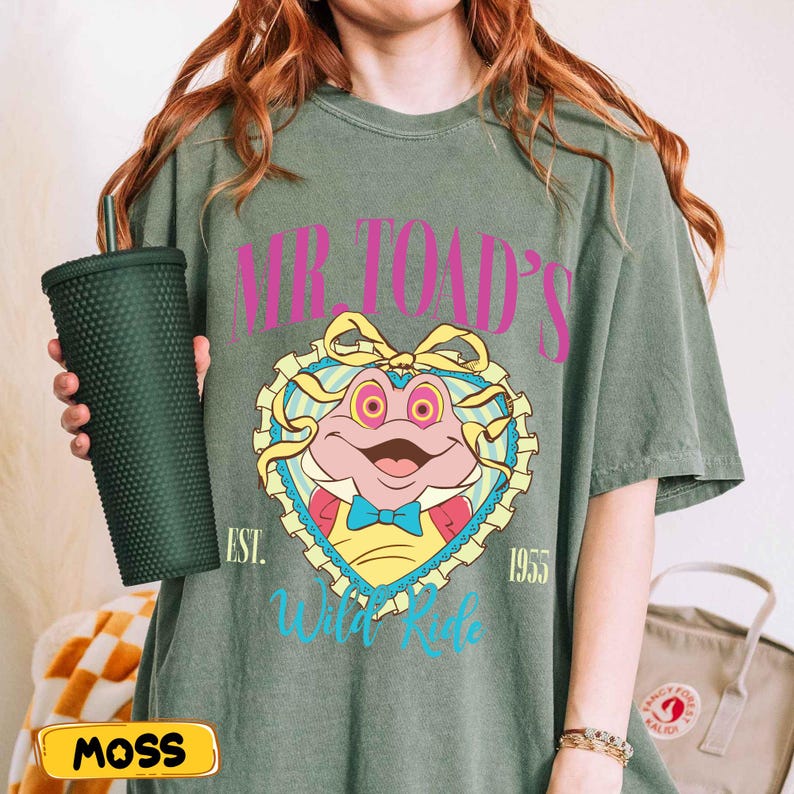 May include: Olive green t-shirt featuring a cartoon frog design with the text "Mr. Toad's Wild Ride" and the year "1955". A dark green studded tumbler is held in the left hand. The word "MOSS" is on a yellow label.