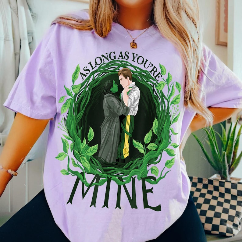 May include: A lilac t-shirt with a graphic of two figures embracing, framed by a green leafy wreath. The text "AS LONG AS YOU'RE" curves above the figures, with "MINE" below. The design is centrally placed on the shirt.