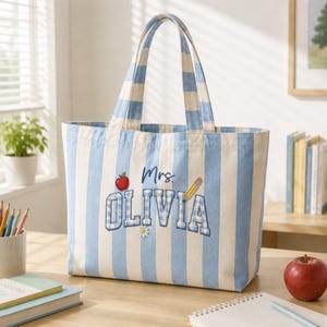 May include: A light blue and white vertical striped tote bag featuring the embroidered text Mrs. OLIVIA in a block font. The design includes a small red apple, a yellow pencil, and a white daisy flower accenting the personalized name.