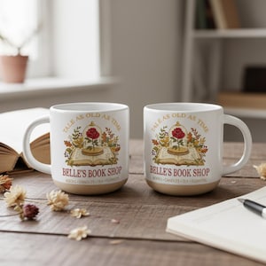 Pode incluir: Duas canecas de cerâmica branca com base bege, cada uma com o texto "TALE AS OLD AS TIME" e "BELLE'S BOOK SHOP". O design inclui uma rosa vermelha numa cúpula de vidro, um livro aberto e folhas de outono. As canecas estão numa superfície de madeira.