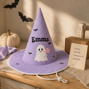 May include: A quilted lavender witch hat featuring a white ghost graphic holding a pink lollipop, small black bat silhouettes, and the name Emma printed in black. The hat includes a white chin strap with an adjustable toggle closure.